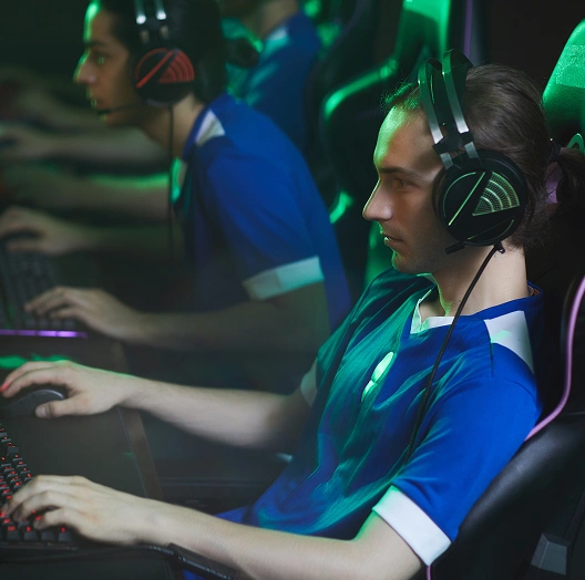 Gamers in blauwe shirts spelen intensief op computers, met een headset op, in een schemerige ruimte met groene verlichting, wat hun concentratie en teamwork benadrukt